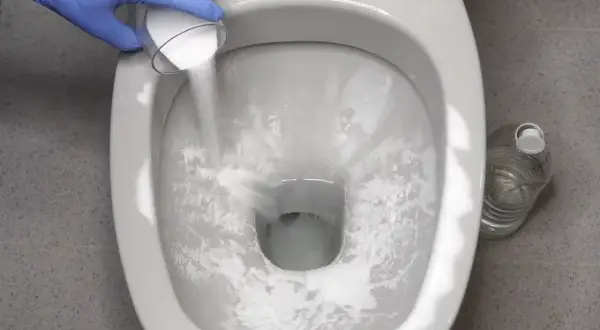 Guanto blu versa polvere bianca nel WC per disincrostare. Bottiglia d'acqua sullo sfondo.