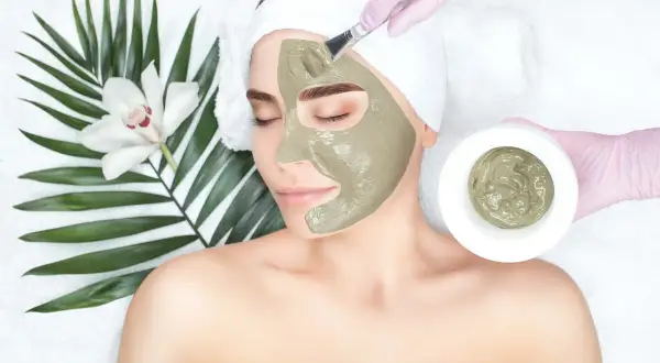 Una donna riceve un trattamento viso con maschera all'argilla verde, applicata con un pennello. Accanto, una foglia di palma e un fiore bianco.