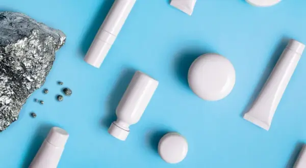 Pietra grezza e piccoli frammenti metallici accanto a flaconi e tubetti bianchi, evocano la purezza dei cosmetici nichel free.