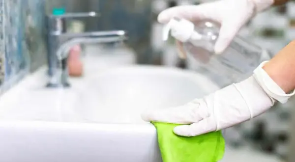 Mani guantate puliscono il lavandino con un panno verde e uno spray, mostrando come pulire il bagno in modo naturale.