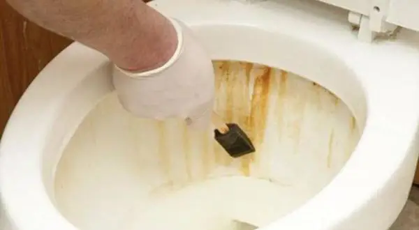 Mano con guanto bianco che usa una spugnetta per rimuovere la ruggine dal water.