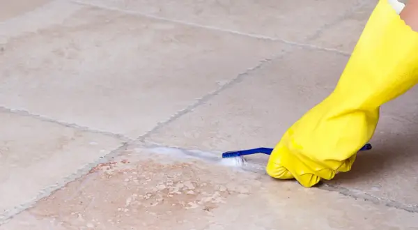 Mano con guanto giallo pulisce le fughe dei pavimenti con uno spazzolino e schiuma.