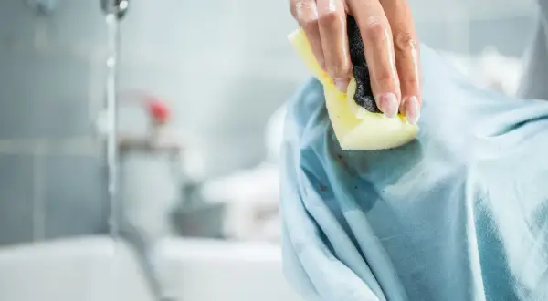 Mano che pulisce una macchia scura su un vestito azzurro con una spugna. Le macchie di profumo vanno via con un po' di pazienza.