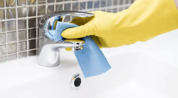 Mano con guanto giallo pulisce rubinetto cromato con panno blu. Come togliere le pietre di sole dal bagno.