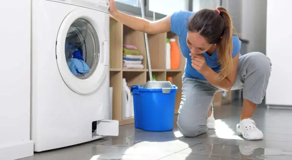 Donna preoccupata guarda la lavatrice che perde acqua dalla vaschetta del detersivo. Sul pavimento si vede una pozza d'acqua.