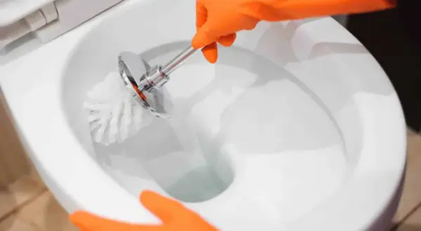 Mani con guanti arancioni puliscono il water, rimuovendo le incrostazioni d'acqua con uno scopino.