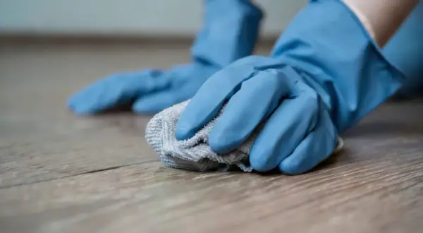 Mani con guanti blu puliscono un pavimento in legno con un panno, creando un detersivo pavimenti fai da te.