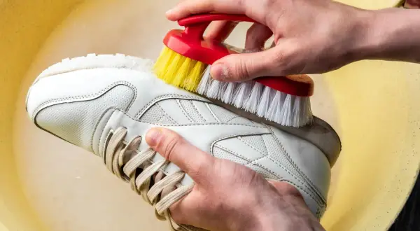 Mani che lavano scarpe di tela bianche a mano con una spazzola in una bacinella gialla.