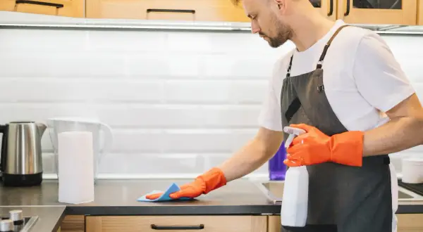 Uomo con guanti arancioni e grembiule pulisce il piano della cucina con uno spray. Ecco come pulire la cucina in modo efficace.
