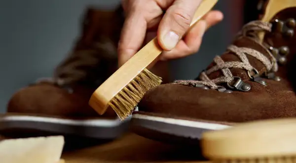 Mano che spazzola una scarpa scamosciata marrone con una spazzola metallica, mostrando come pulire la pelle scamosciata delle scarpe per mantenerla in forma.
