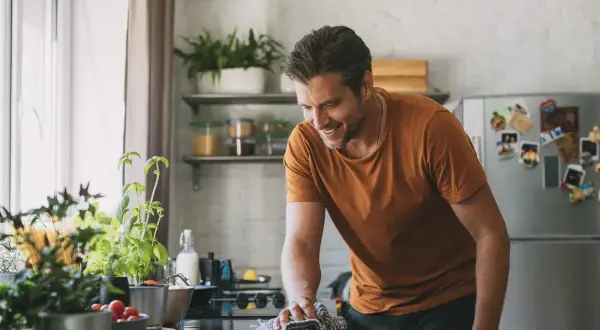 Uomo sorridente pulisce il piano cottura con un panno, mentre sul bancone ci sono piante aromatiche e pomodorini. Forse usa cristalli di soda per lucidare.