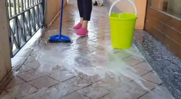 Pulizia del terrazzo esterno: una scopa blu lava le piastrelle con acqua saponata, accanto un secchio giallo.
