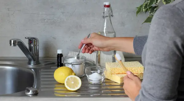 Mano che preleva bicarbonato da un barattolo, con limone, percarbonato e spugne. Differenza tra percarbonato e bicarbonato per pulizie ecologiche.