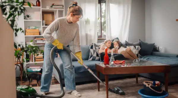 Donna con guanti gialli aspira il tappeto, mentre due bambine guardano il telefono sul divano. Una guida pratica su come pulire una casa sporchissima.