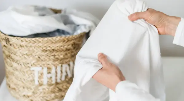 Mani che piegano un lenzuolo bianco, vicino a un cesto di vimini con biancheria. Speriamo non ci siano macchie candeggina!