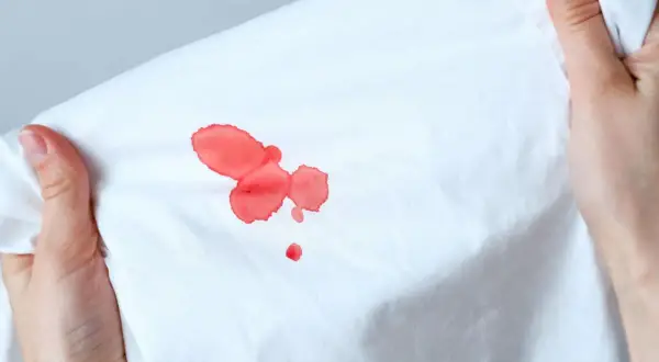 Mani che tengono un tessuto bianco con macchie di sangue rosso vivo. Scopri come togliere le macchie di sangue in modo efficace.