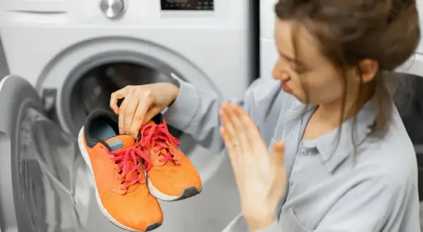 Donna con espressione disgustata tiene in mano scarpe da ginnastica arancioni, pronte per essere lavate in lavatrice.