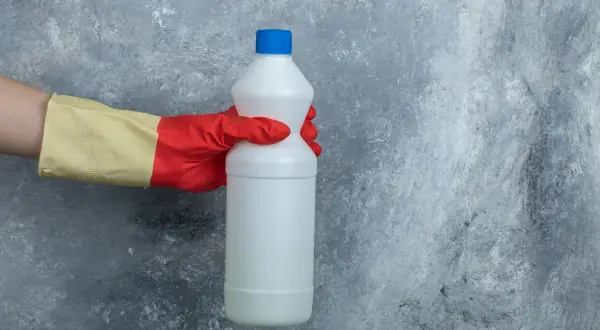 Mano con guanto rosso e giallo tiene una bottiglia di candeggina, pronta per la lavatrice.