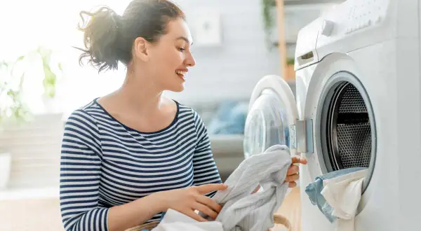 Donna sorridente carica la lavatrice per il lavaggio quotidiano. Un cesto di panni è accanto alla lavatrice aperta.