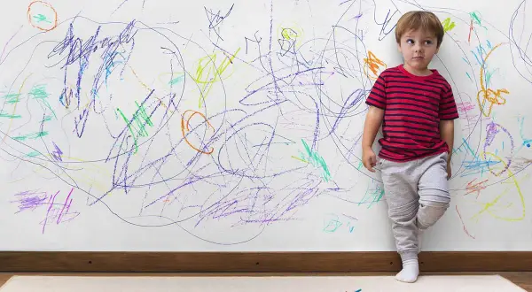 Bambino vicino a un muro pieno di scarabocchi colorati. Come togliere macchie dal muro colorato dopo il suo capolavoro.