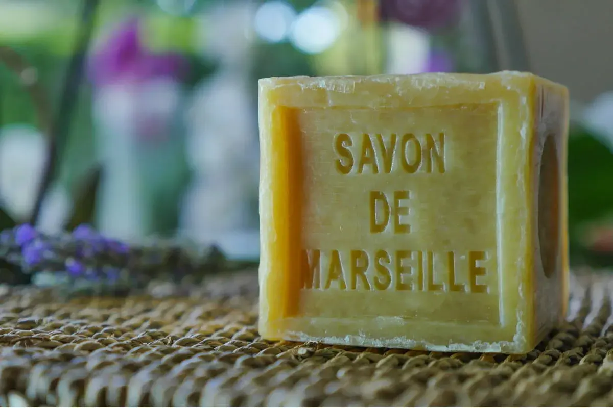 Un pezzo di sapone bucato, un classico sapone di Marsiglia, su una trama di paglia. Sullo sfondo, lavanda e fiori sfocati.