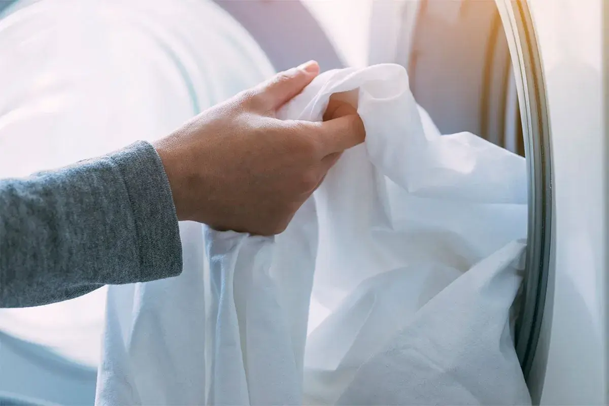 Mano che inserisce un lenzuolo bianco nella lavatrice. Ricorda che candeggina e bicarbonato si possono mischiare con cautela per un bucato più bianco.