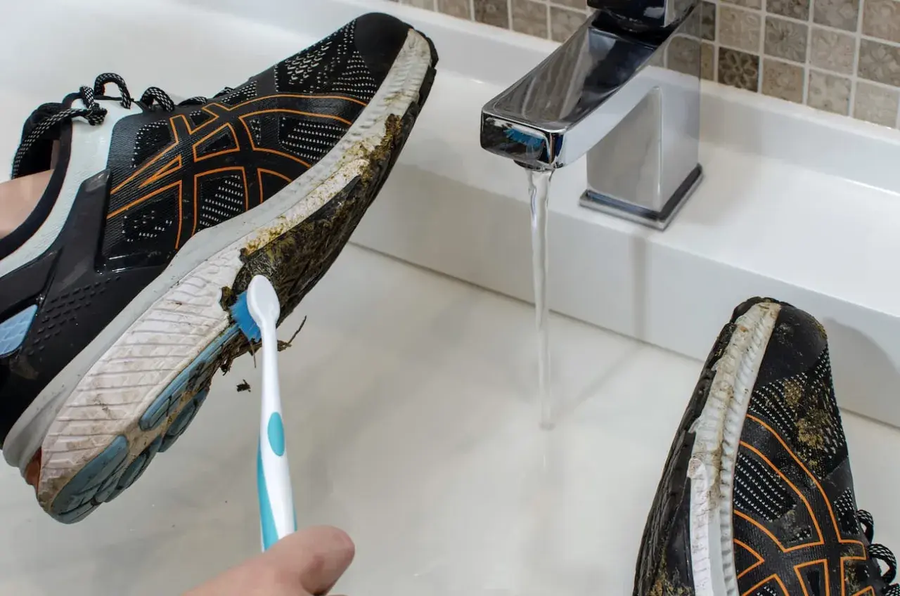 Come lavare le scarpe da running: una mano con uno spazzolino pulisce il fango dalla suola di una scarpa sotto un rubinetto aperto.