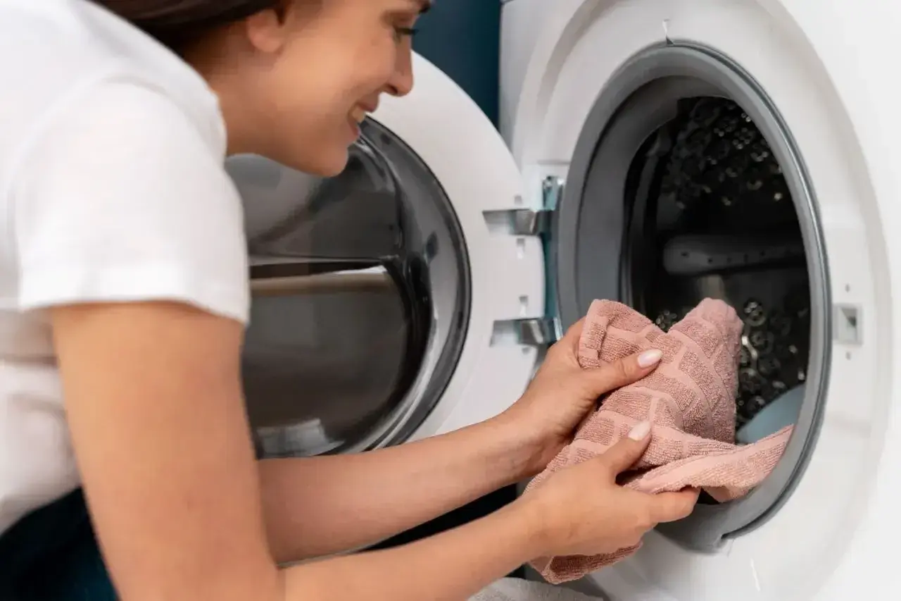 Donna sorride mentre mette un asciugamano rosa nella lavatrice, pronta per un bucato fresco.