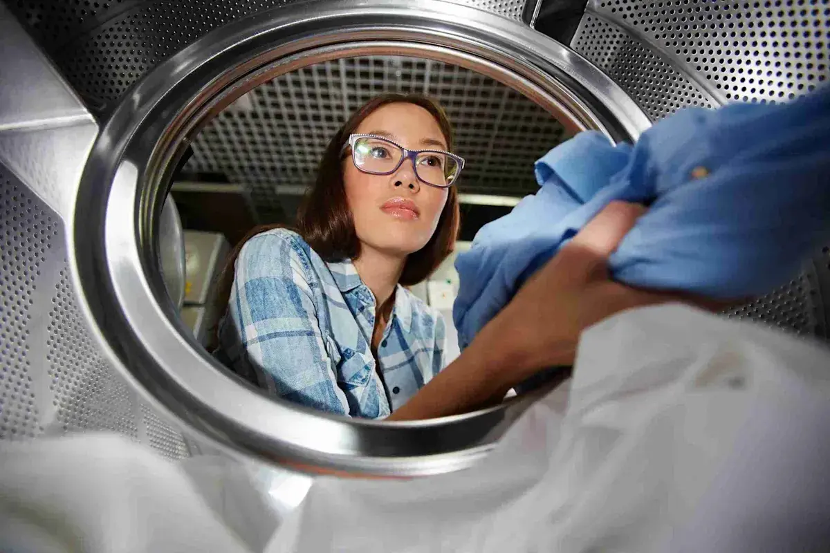 Donna con occhiali guarda dentro cestello lavatrice, mentre una mano inserisce un capo blu. Utile per capire come pulire cestello lavatrice.