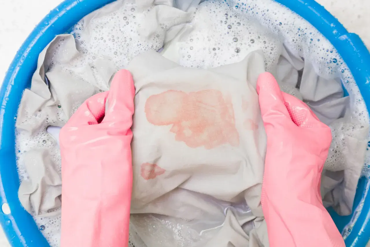Mani con guanti rosa che lavano un vestito macchiato di rosso in una bacinella blu con acqua saponata. Ecco come sbiancare i vestiti che hanno preso colore.