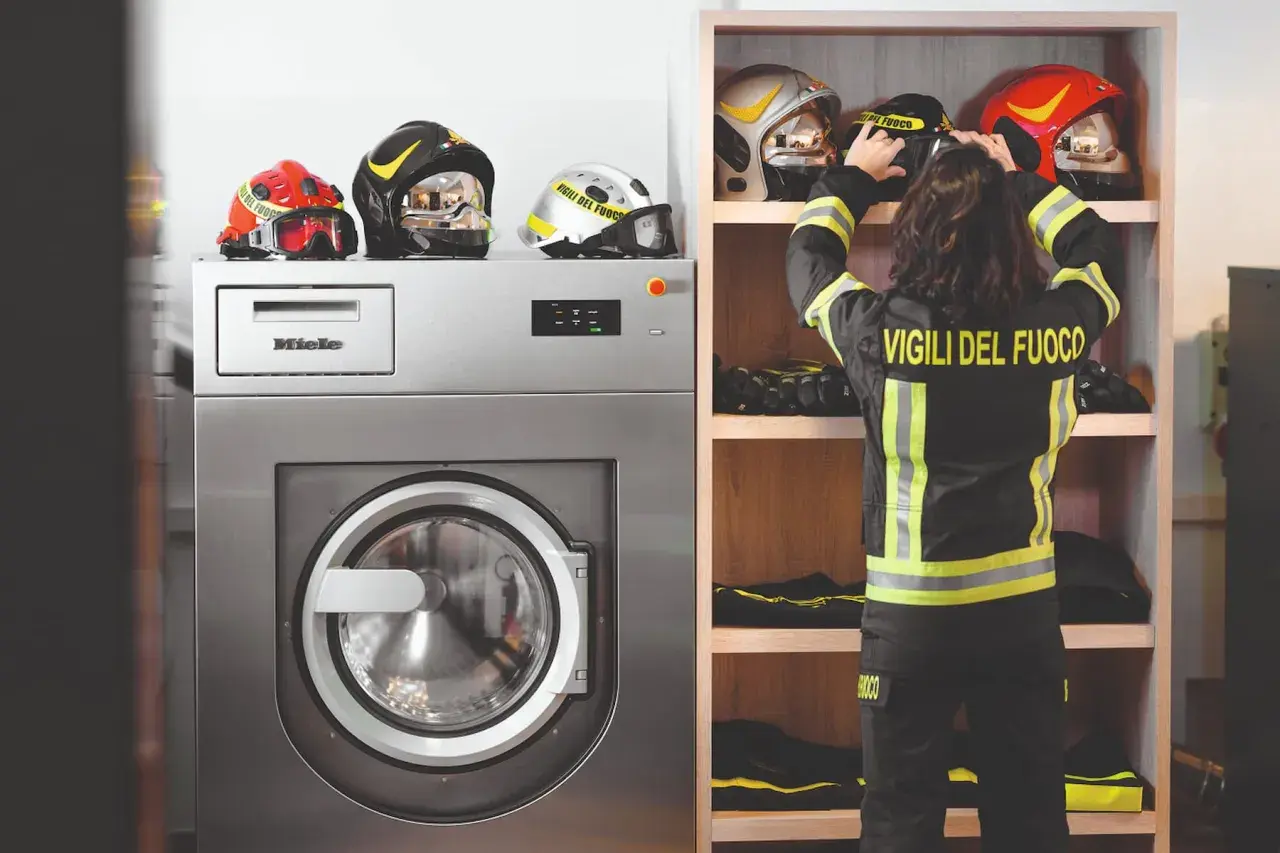 Vigile del Fuoco sistema i caschi accanto a una lavatrice industriale, pronta per il lavaggio abiti da lavoro.