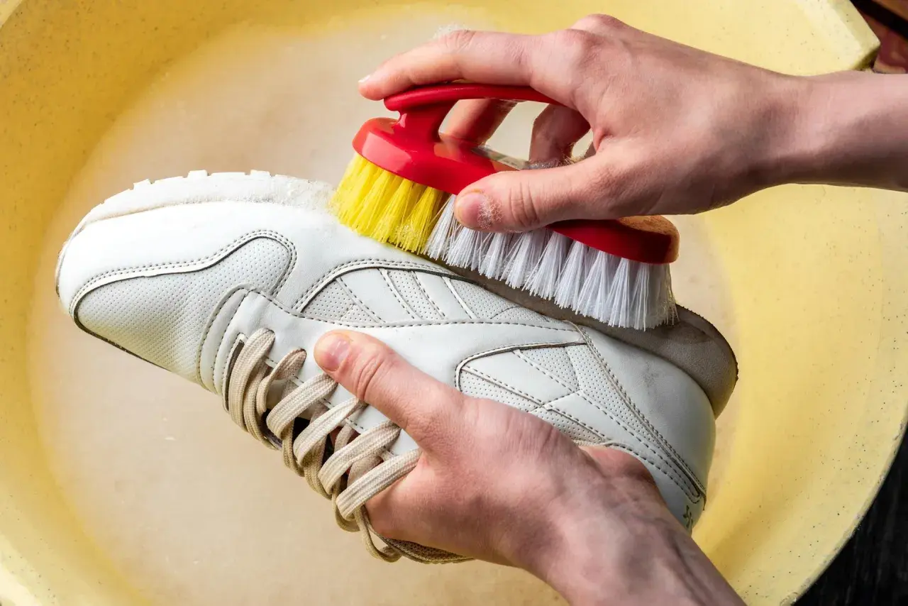 Mani che lavano scarpe di tela bianche a mano con una spazzola in una bacinella gialla.
