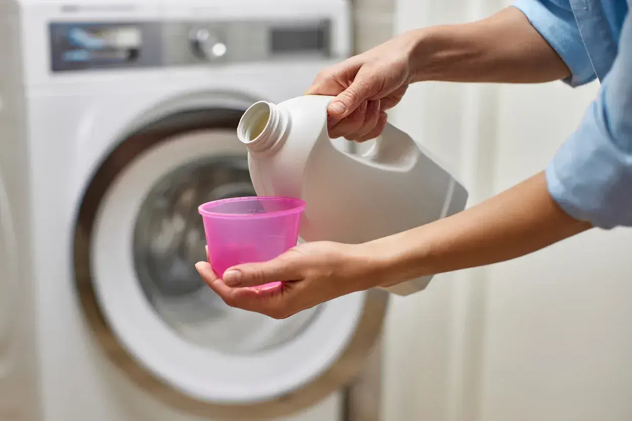 Mani versano candeggina in un misurino rosa, vicino a una lavatrice. Quanto tempo ammollo candeggina?