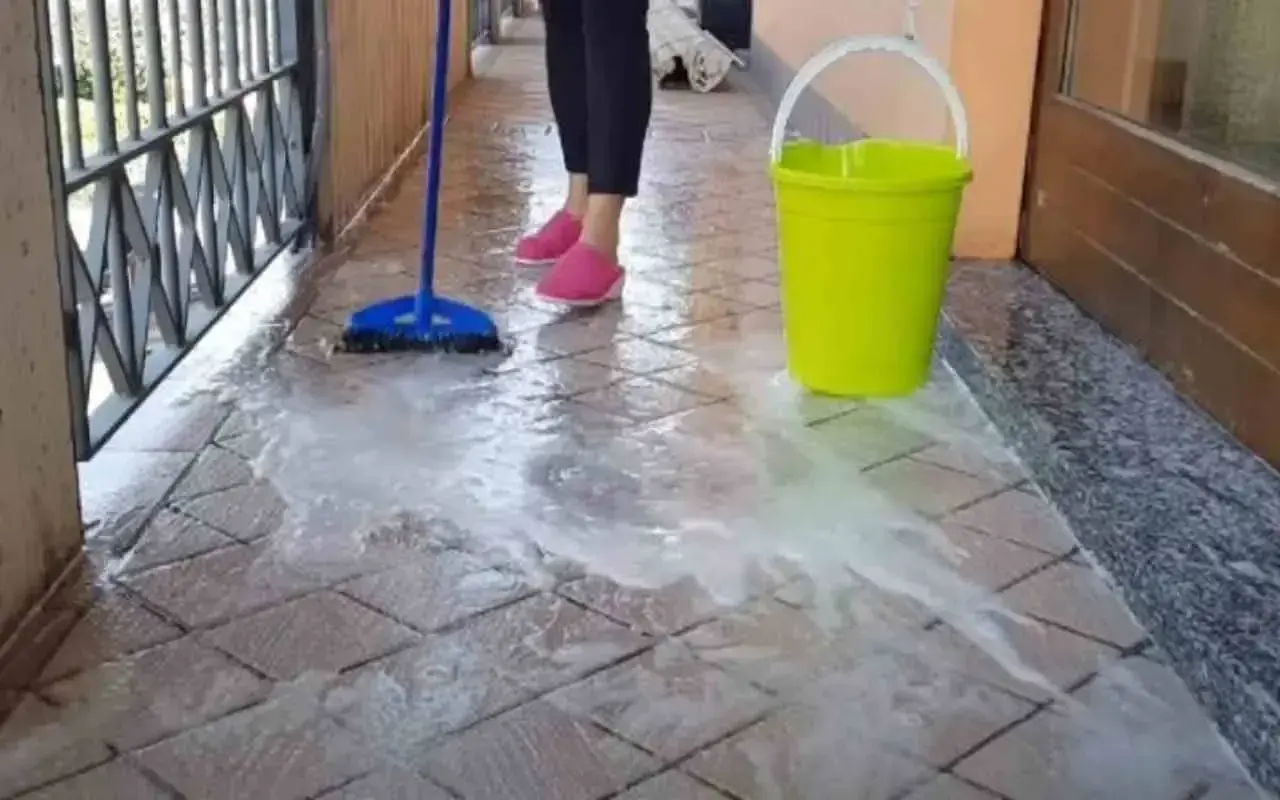 Pulizia del terrazzo esterno: una scopa blu lava le piastrelle con acqua saponata, accanto un secchio giallo.