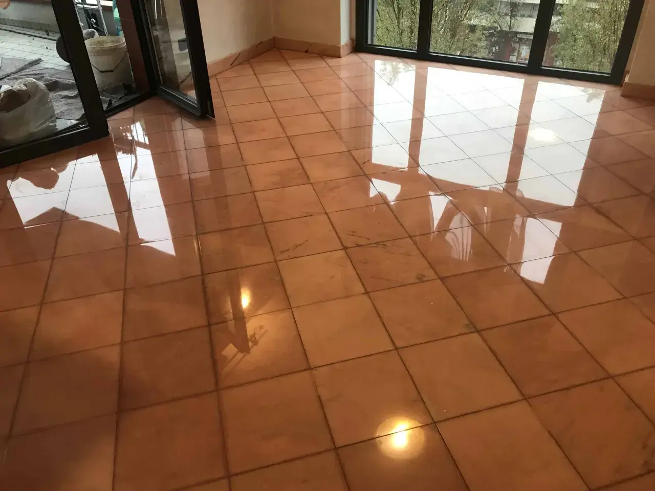 Pavimento in cotto lucido, riflessi di luce e finestre. Si intravede un secchio e sacchi, forse per pulire marmo con aceto.