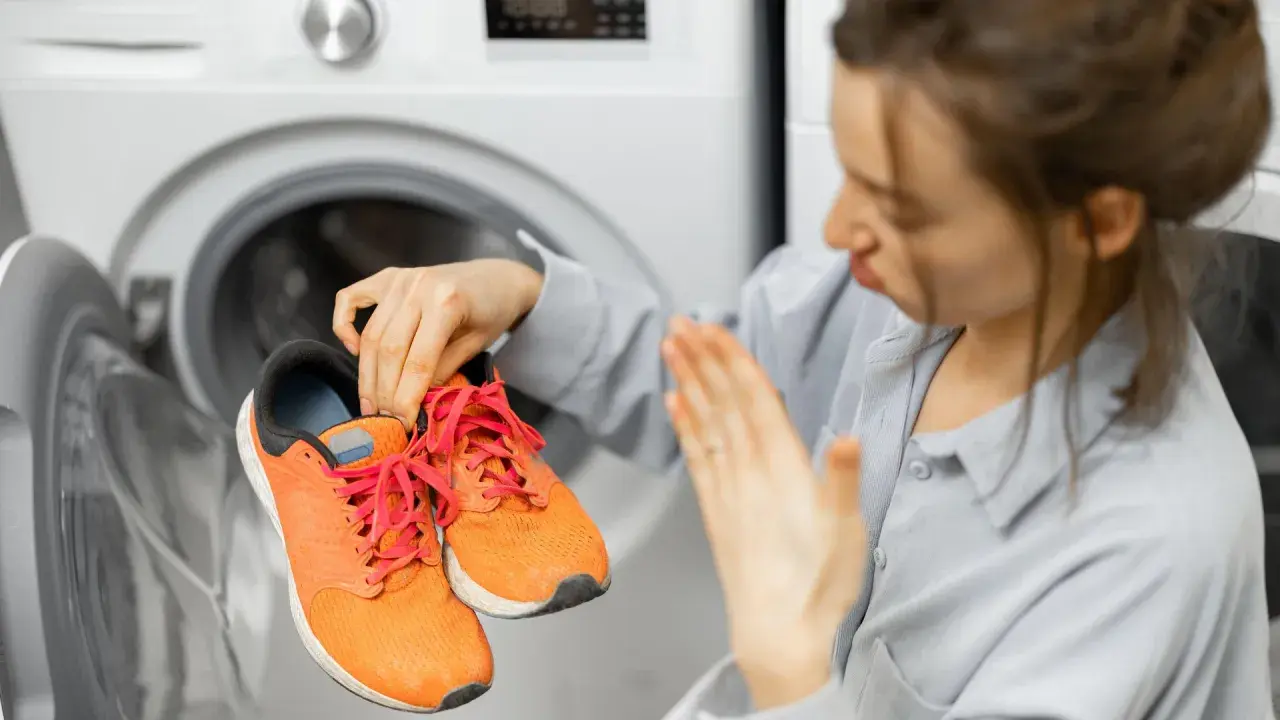 Donna con espressione disgustata tiene in mano scarpe da ginnastica arancioni, pronte per essere lavate in lavatrice.