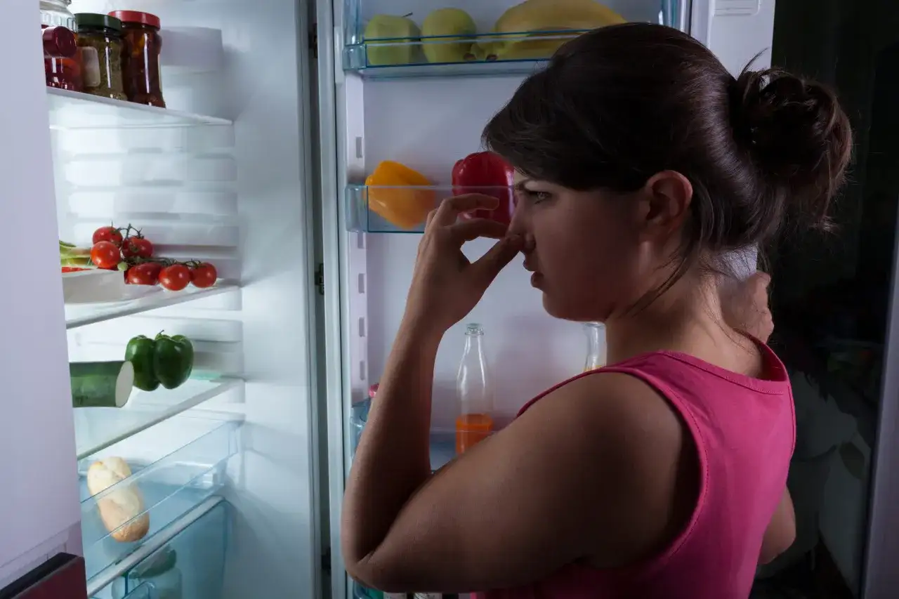 Donna con mano sul naso davanti a un frigorifero pieno di cibo, preoccupata per come togliere la puzza dal frigo.