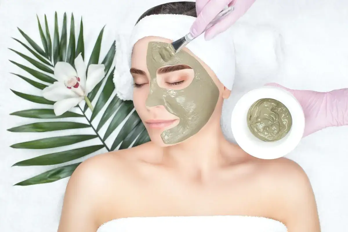 Una donna riceve un trattamento viso con maschera all'argilla verde, applicata con un pennello. Accanto, una foglia di palma e un fiore bianco.