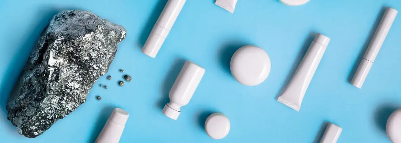 Pietra grezza e piccoli frammenti metallici accanto a flaconi e tubetti bianchi, evocano la purezza dei cosmetici nichel free.