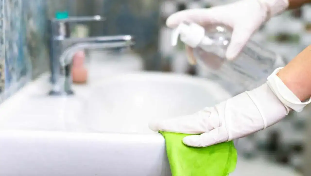 Mani guantate puliscono il lavandino con un panno verde e uno spray, mostrando come pulire il bagno in modo naturale.