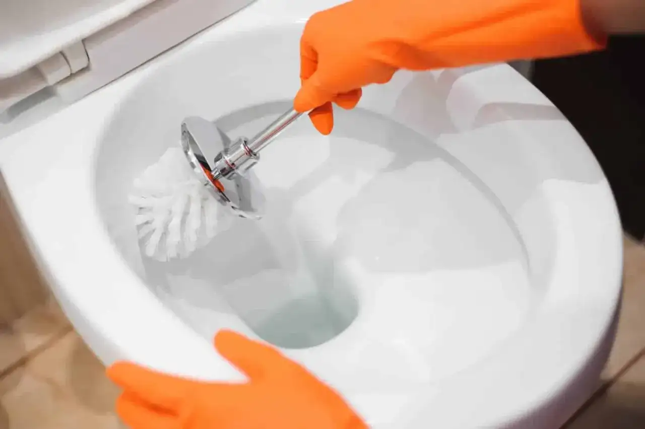 Mani con guanti arancioni puliscono il water, rimuovendo le incrostazioni d'acqua con uno scopino.