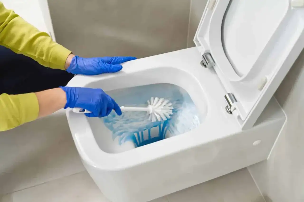 Mani con guanti blu puliscono il water, creando schiuma e macchie blu nel water con uno scopino.