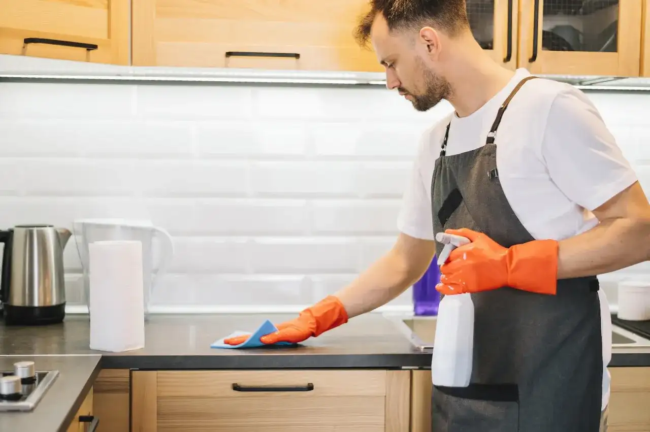 Uomo con guanti arancioni e grembiule pulisce il piano della cucina con uno spray. Ecco come pulire la cucina in modo efficace.