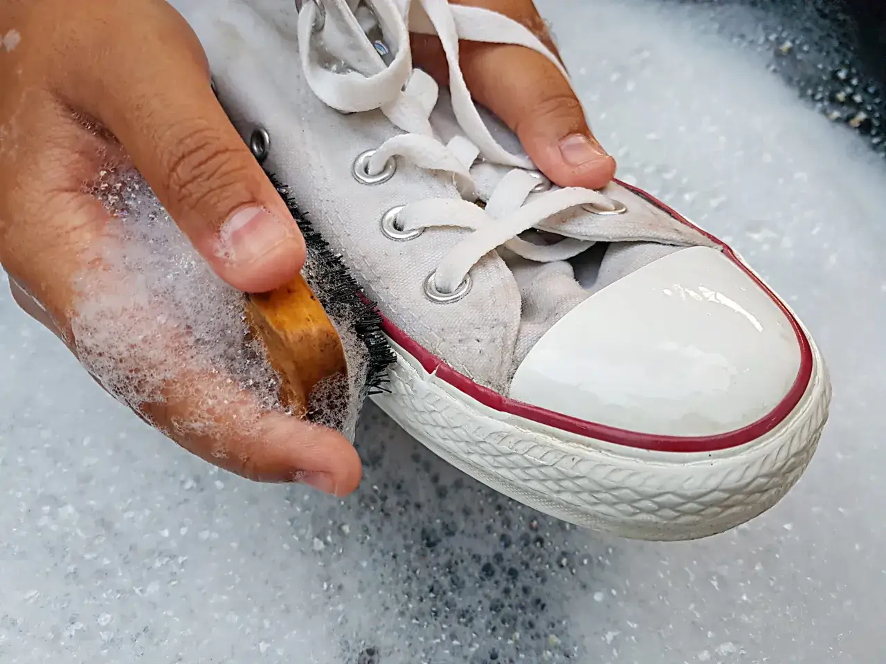Mani che lavano una scarpa di tela bianca con una spazzola e sapone, mostrando come lavare le scarpe di tela a mano per farle tornare come nuove.