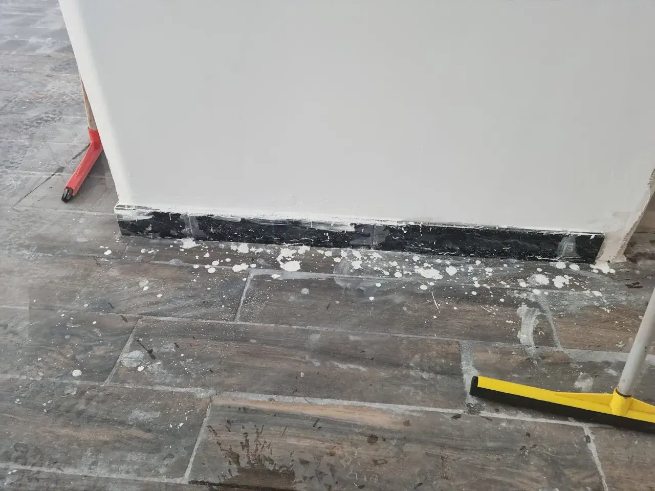 Rimozione vernice dal marmo: gocce bianche sul battiscopa nero e sul pavimento in finto legno, con scopettone giallo.