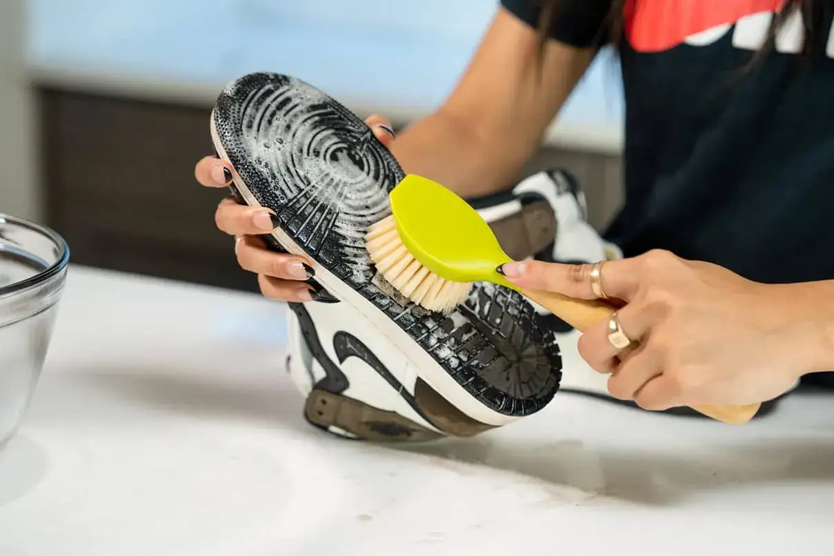 Mani che puliscono la suola di una scarpa con una spazzola gialla e sapone, mostrando come lavare le scarpe per farle tornare come nuove.