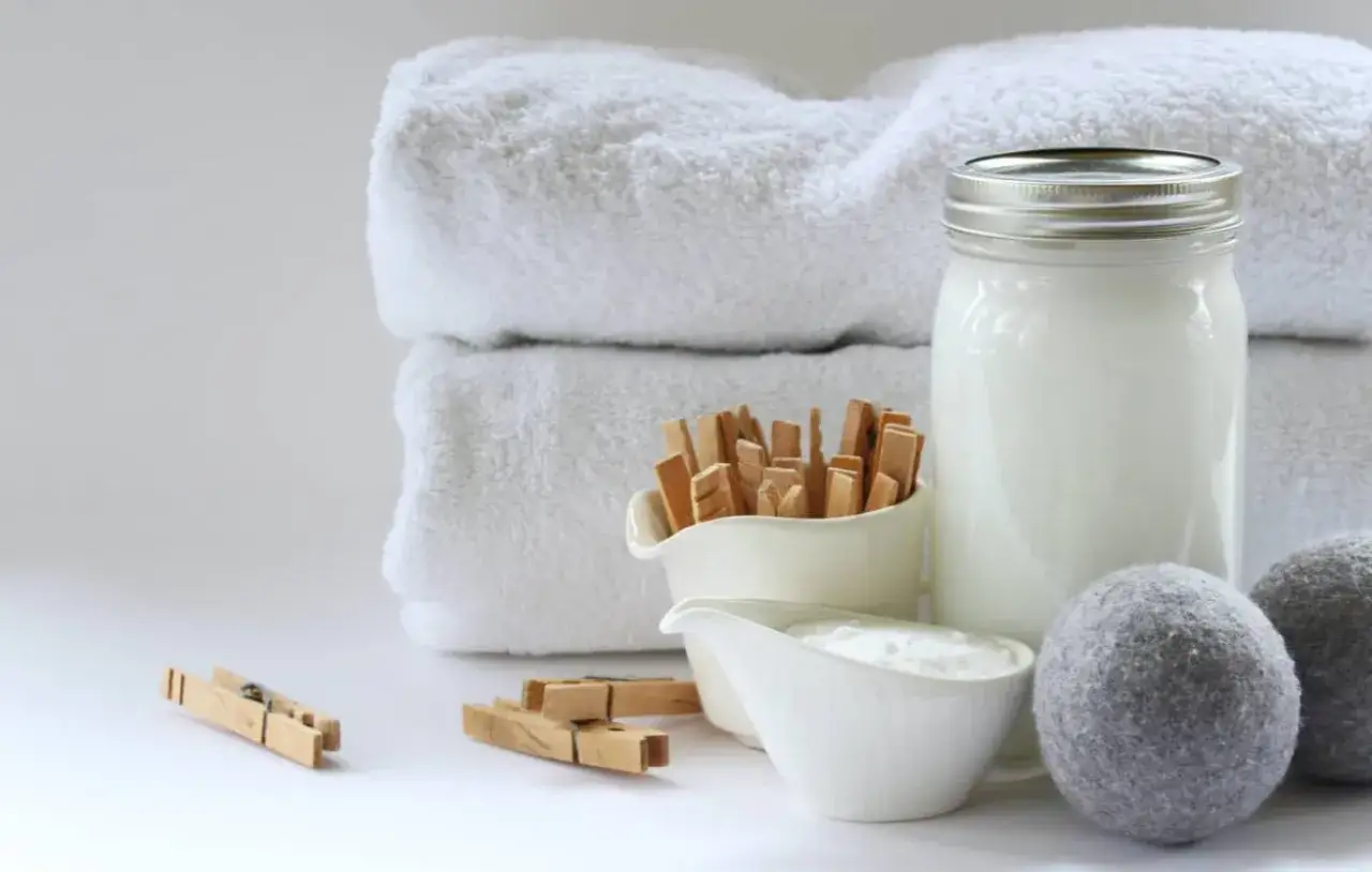 Panni bianchi morbidi, un barattolo di vetro con liquido bianco, ciotole con ingredienti e mollette di legno: tutto per un ammorbidente naturale.