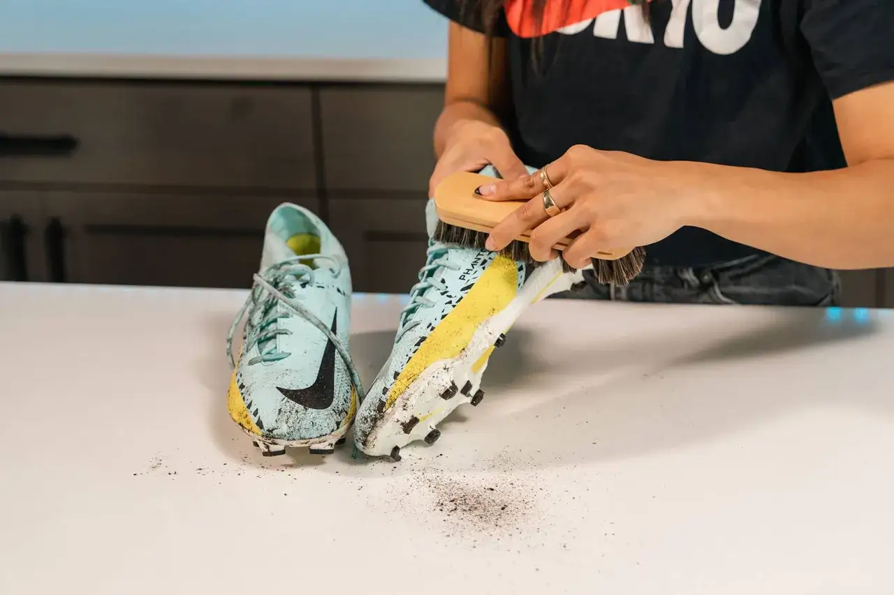 Mani che puliscono scarpe da calcio con una spazzola, mostrando come lavare le scarpe da calcio dopo una partita fangosa.