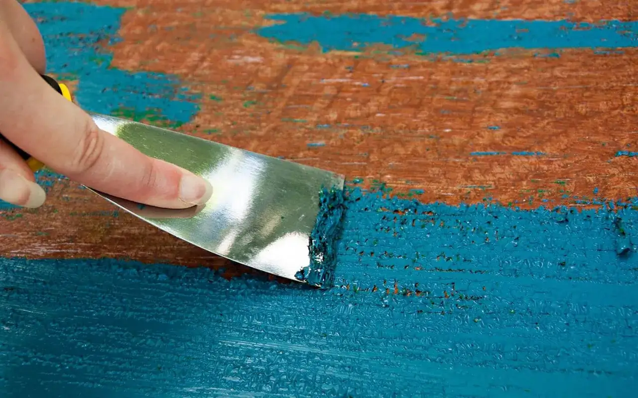 Una mano raschia via una macchia di pittura blu da una superficie di legno grezzo.