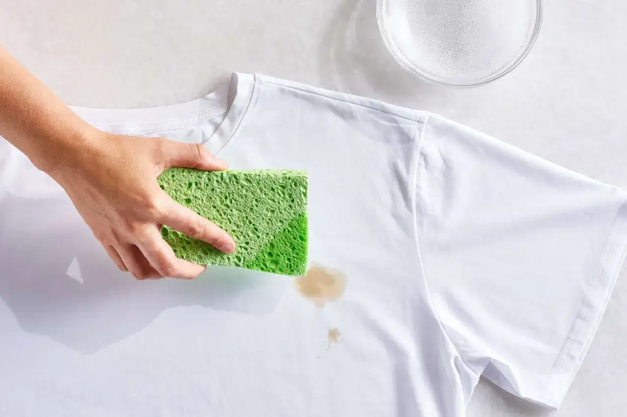 Mano che usa una spugna verde per smacchiare una maglietta bianca macchiata.